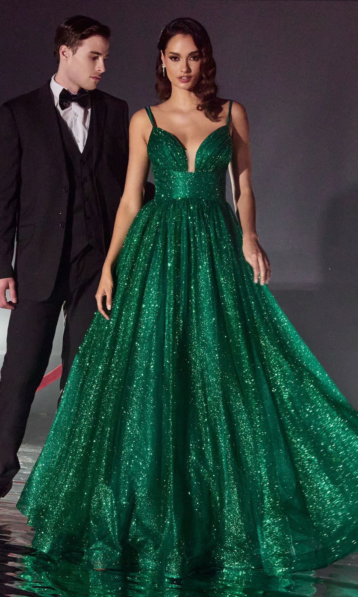 A woman in the Corset-Back Long Glitter Prom Ball Gown CD996 stands in front of a man in a black tuxedo against a dark background.